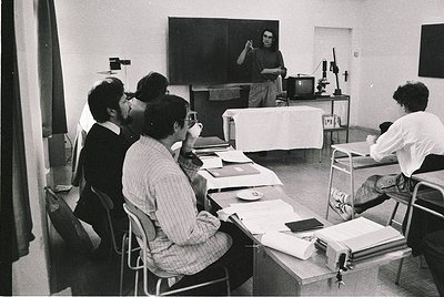 Black-and-white classroom scene featuring a lecturer gesturing at a chalkboard while students take notes, 1960s–1970s. Wooden...