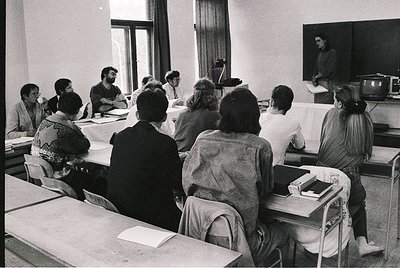 Black-and-white classroom scene with 12 seated students facing a lecturer at a podium. Wooden desks, vintage chairs, and a TV...