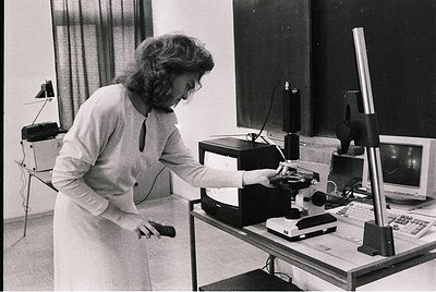 A professional woman in a lab coat operates vintage optical equipment, likely a microscope or slide projector, in a 1970s-era...