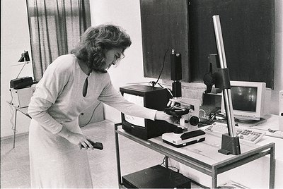 Woman in 1970s lab coat operates vintage slide projector with overhead lamp, surrounded by analog equipment. Industrial light...