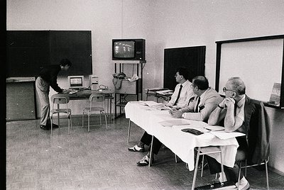 Vintage black-and-white office scene with four men in formal attire (1960s-70s). One man stands at a typewriter, others seate...