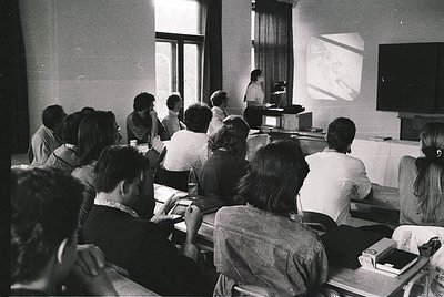 Black-and-white lecture hall scene from the 1960s–70s, featuring a speaker at a podium with projected slides. Audience faces ...