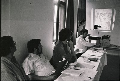 Mid-20th century conference room with men in formal attire (1960s–1970s). Projector displays a detailed map or diagram on a w...