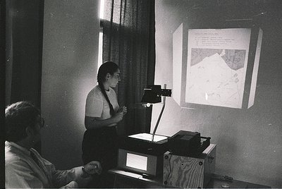 Mid-20th century classroom lecture on geography or cartography. Woman in 1960s-style attire projects a detailed map onto a wa...