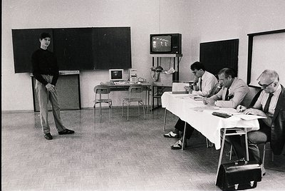 Black-and-white 1970s office scene: a man in a sweater stands beside a chalkboard, while four seated professionals review doc...