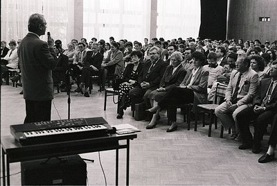 Black-and-white lecture hall scene featuring a speaker addressing a seated audience of ~80 individuals in formal attire (1960...