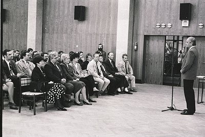 A formal indoor gathering in a mid-century conference hall, featuring a speaker addressing an attentive audience seated in wo...