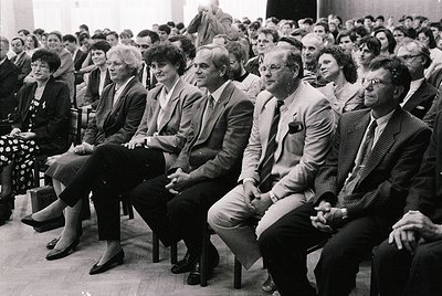 1970s indoor gathering of formal attire attendees in a packed auditorium. Wooden chairs, tiered seating, and a stage backdrop...