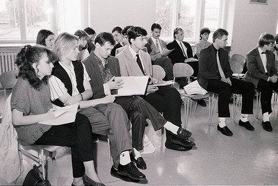 Classroom setting featuring 1970s-era students in formal attire—mixed-gender group seated on plastic chairs, reading papers o...