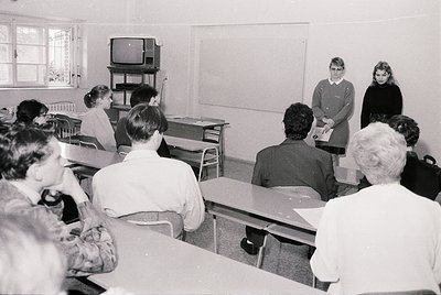 Mid-20th century classroom with vintage desks, blackboard, and CRT TV. Two teachers (one in uniform) and students seated/stan...