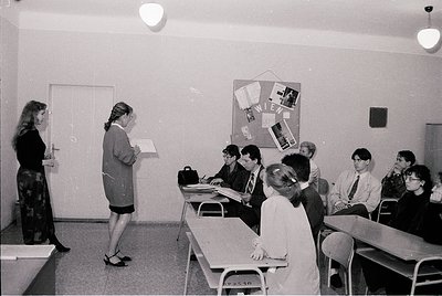 Spacious classroom with 1970s/80s design—teacher in skirt suit directs students seated at desks. Bulletin board displays "VIE...