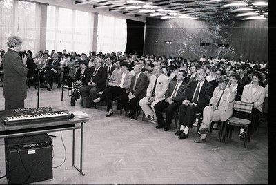 A black-and-white lecture hall scene from the 1970s–1980s, featuring a speaker at a podium with a vintage keyboard and amplif...