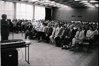 Mid-century lecture hall with tiered seating, featuring a speaker at a podium with a keyboard instrument (likely a Wurlitzer)...
