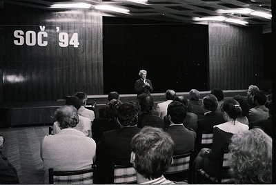 Black-and-white lecture hall shot at the 1994 Summer Olympics (SOC) in Lillehammer, Norway. Speaker addresses seated audience...