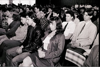 Black-and-white shot of a packed indoor lecture hall, likely from the **1970s-1980s**. Audience of young adults in formal att...