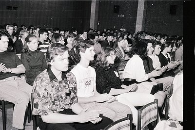 Black-and-white indoor lecture hall filled with seated students, likely 1970s–1980s. Wooden benches and tiered seating create...