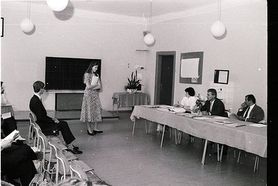 Mid-20th century classroom/conference setting with vintage 1960s-70s decor. Woman in floral dress addresses seated men at a l...
