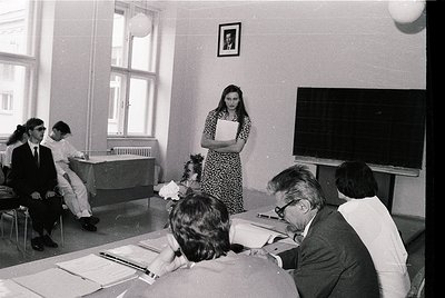 Mid-20th century classroom setting with formal attire and vintage teaching aids. A woman in a patterned dress stands at a cha...