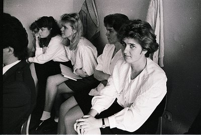 Group of four women in 1960s-era formal attire—blouses, jackets, and short hair—sitting in a dimly lit indoor setting, possib...