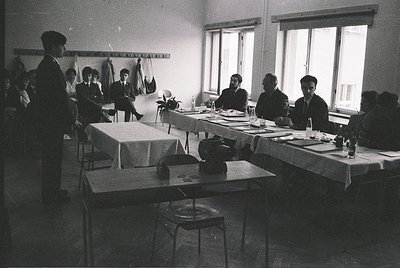 Classroom setting with vintage typewriters, likely mid-20th century. Six seated students and a teacher at desks with white ta...