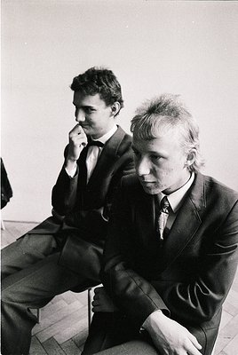 Two men in formal attire sit on stools in an indoor setting, likely a 1970s European office or meeting space. The man on the ...