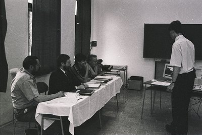 Black-and-white office scene from the 1970s–1980s, featuring five men in formal attire seated around a table with documents. ...