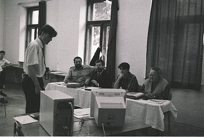 Retro office scene featuring early desktop computers (1980s-era) in a classroom-like setting. Six individuals engage with ter...