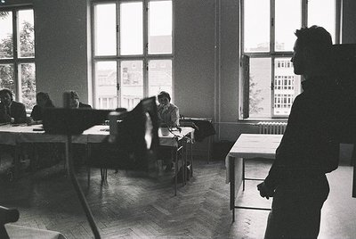 Classroom setting with mid-century industrial design—wooden desks, metal chairs, and large windows revealing urban buildings....
