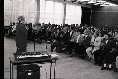 A speaker addresses a packed indoor audience in a formal setting, likely a conference or lecture hall. The speaker stands at ...
