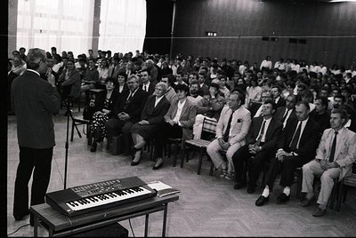 Black-and-white lecture hall scene from the 1960s–70s, featuring a speaker at a podium with a keyboard instrument, likely a m...