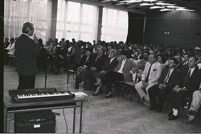 A formal indoor lecture or conference in a mid-20th-century setting, featuring a speaker at a podium with a keyboard instrume...