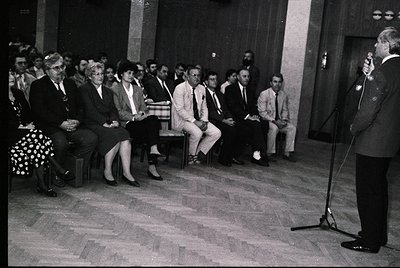 Black-and-white indoor gathering in a formal setting, likely mid-20th century. A speaker stands at a podium with a microphone...