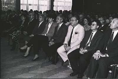Mid-20th century formal gathering in a tiered auditorium. Attendees in suits, ties, and dresses sit attentively, suggesting a...