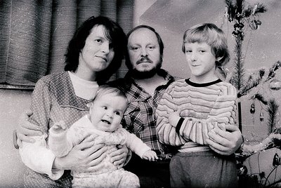 Family portrait from the 1970s–1980s, featuring three adults and an infant. Mother holds baby in a knitted sweater, father we...