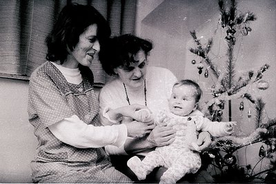 Two women cradle a baby in a festive indoor setting, likely a 1960s-1970s Christmas scene. The woman on the left wears a patt...