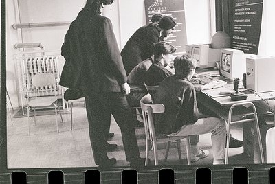 Black-and-white classroom scene featuring early computer education. Three students seated at desks with CRT monitors, likely ...