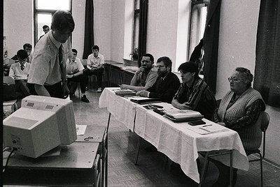 Mid-20th century classroom setting with early computer demo. Man in striped shirt operates a bulky desktop computer (1970s-80...