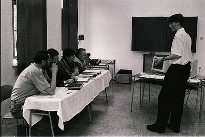 Mid-20th century conference room with four seated attendees reviewing documents at a table, one standing presenter using a pr...