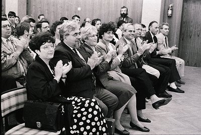 Audience clapping during a formal event in the 1970s, likely a conference or lecture. Indoor setting with wooden chairs and a...