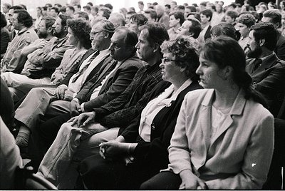 Black-and-white audience of seated adults in formal attire, likely attending a public event or lecture. Men wear suits, ties,...