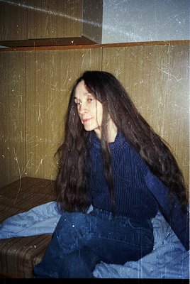 Portrait of a person with long, straight hair in a vintage-style sweater and cropped trousers, seated on a bed with a striped...