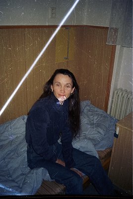 Vintage indoor portrait of a woman seated on a single bed in a utilitarian room, likely from the 1970s–1980s. She wears a dar...