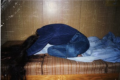 Person resting head on a wooden bench in an indoor setting, likely a public transport station or waiting area. The worn, stri...