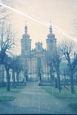 Baroque-style church with twin domed towers and ornate façade, framed by bare winter trees. Symmetrical clock placement on ce...