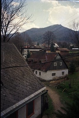 Rustic alpine village with timber-framed houses and stone roofs, framed by bare winter trees. Snow-capped mountains loom in t...
