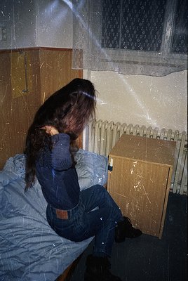 Vintage-style interior shot of a person sitting on a bed with a radiator beside them. The individual wears a long-sleeve shir...