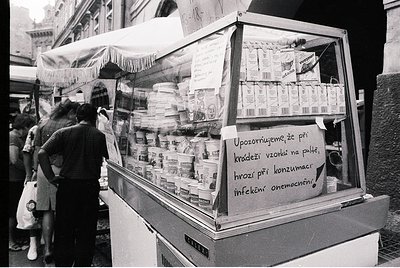 Street-side vending cart in black-and-white, displaying packaged dairy products (milk, yogurt) with handwritten warning in Cz...