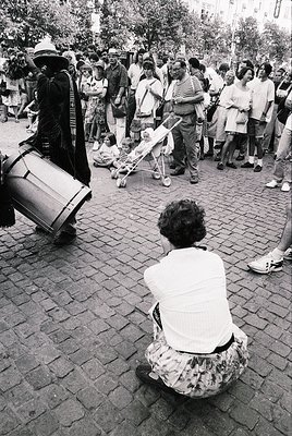 Street musician playing a large frame drum beside a seated woman in floral dress, 1970s urban setting. Crowd of casually dres...