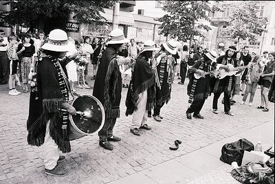 Street performance featuring traditional musicians in folk attire—wide-brimmed hats, layered vests, and fringe—playing drums,...