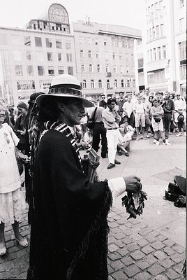 Street musician in traditional attire plays accordion to a gathered crowd in an urban plaza. Architectural details include mi...